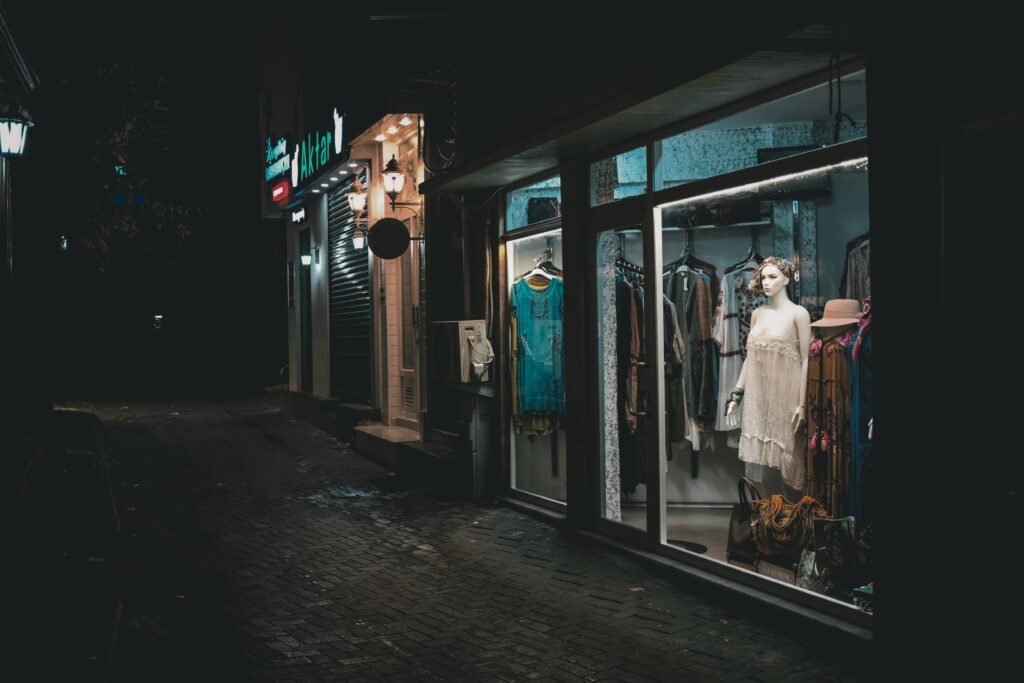 A boutique's fashion display brightly lit in a dark alley at night, showcasing elegant dresses.